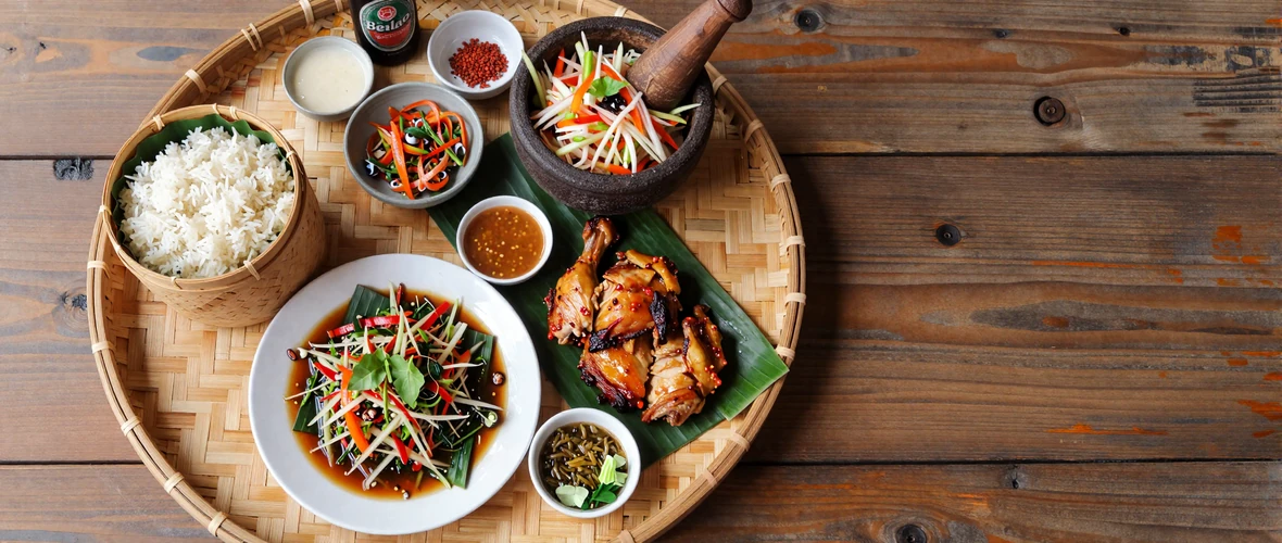 Traditional Lao feast spread with sticky rice, laap salad, papaya salad, grilled chicken, and Beerlao on a bamboo tray